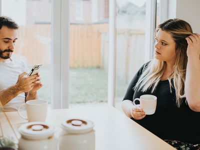 A woman and man sit at a kitchen table. The man looks down at his phone and subsequently ignores the woman. The woman looks insecure and hurt as she looks at him, hoping for some attention. Space for copy.