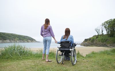 Woman in wheelchair holding hands with woman standing, overlooking a beautiful seaside view
