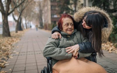 older woman in a wheelchair with red hair being helped by a younger woman, both wearing warm coats