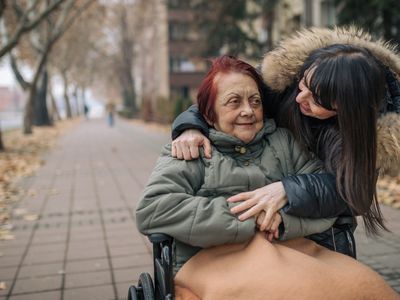 older woman in a wheelchair with red hair being helped by a younger woman, both wearing warm coats