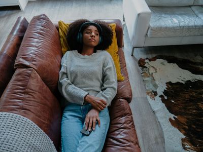 melancholic mixed race female wearing headphones listening to sad moving while holding smartphone lying on leather sofa in modern lounge