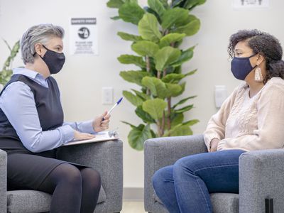 Female therapist listening attentively to a patient during a therapy session, both are wearing protective face masks during COVID-19.