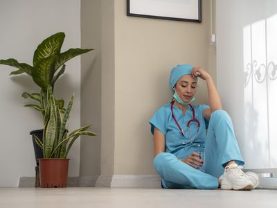 Overworked female medical professional wears face mask, blue uniform and gloves sits on hospital floor
