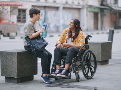 asian chinese mid adult woman talking to her indian female friend with disability on wheelchair at city street sidewalk