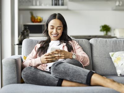 Smiling woman using mobile phone while sitting on sofa. Young female is text messaging in living room. She is relaxing at home.