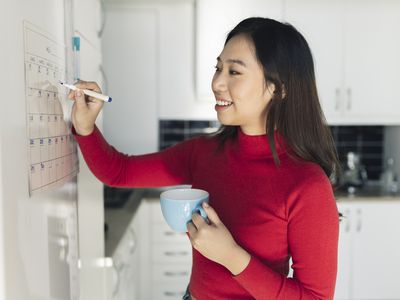 Smiling woman with coffee cup writing on calendar in kitchen