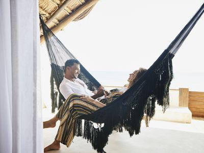 smiling couple holding hands while sitting in hammock on deck of luxury tropical villa