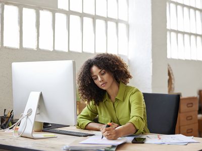 Businesswoman writing on document while sitting at desk. Female professional is working at home office. She has curly hair.