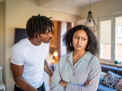 Woman is frowning and looking away from her boyfriend with her arms crossed, as he is looking worriedly into her face as they stand at home in their lounge