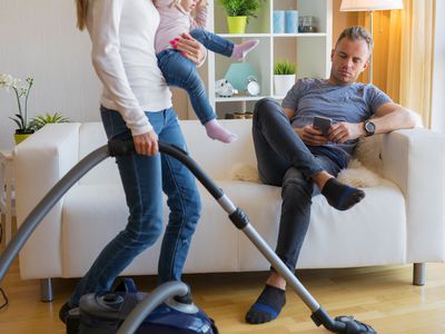 Woman with small child in her hands doing housekeeping while man sitting on couch and relaxing.