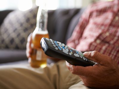 Man Sitting On Sofa Holding TV Remote And Bottle Of