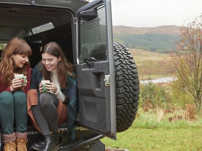 two women sitting in the back of an open car drinking coffee in the autumn