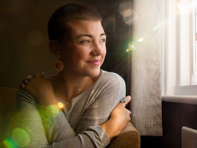 Young woman with short hair and a nose piercing hugs herself and looks out a window