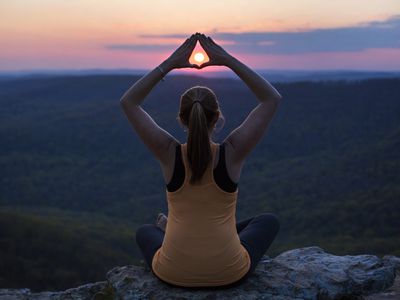 Woman sitting outside in lotus position with the setting sun framed with her hands