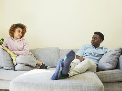Young couple sitting apart on the sofa. They are looking at each other with irritated expressions.